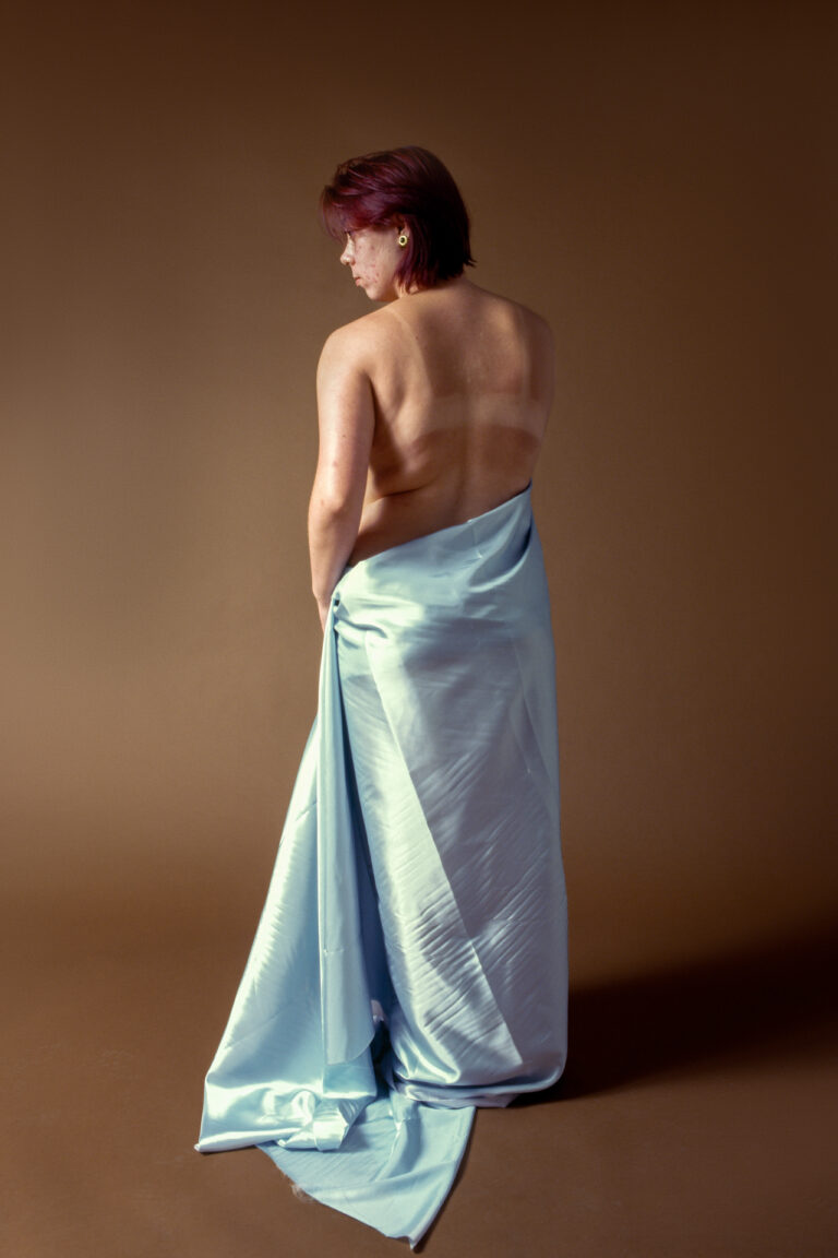 Cut From The Same Cloth An image of a young woman with short red hair stands with her sunburnt back towards the camera, the lower half of her body only covered by a blue sheet.