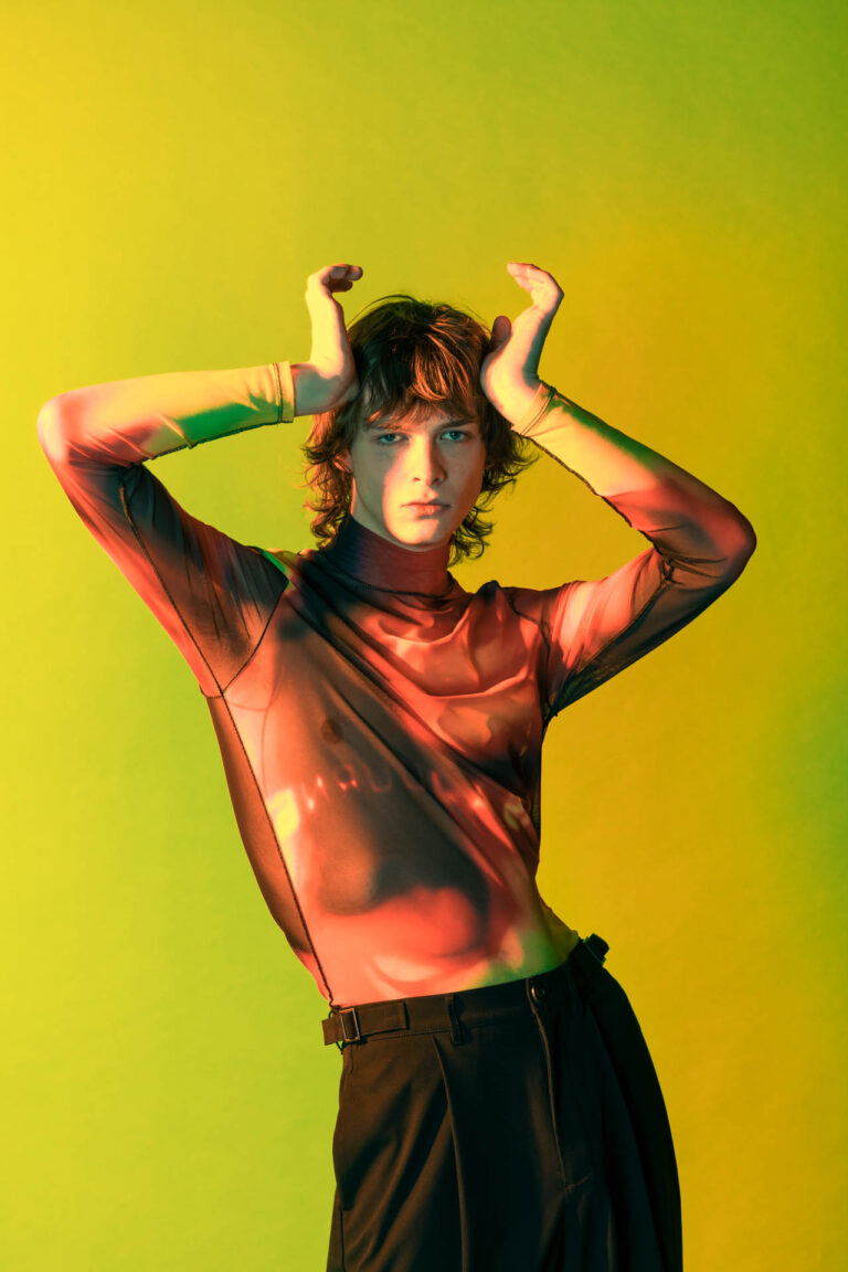 A man wearing black pants and a flame-patterned shirt stands in front of a green background and is lit up through a yellow hue. He is resting his hands on the tip of his forehead as if they are imitating the shape of a crown.