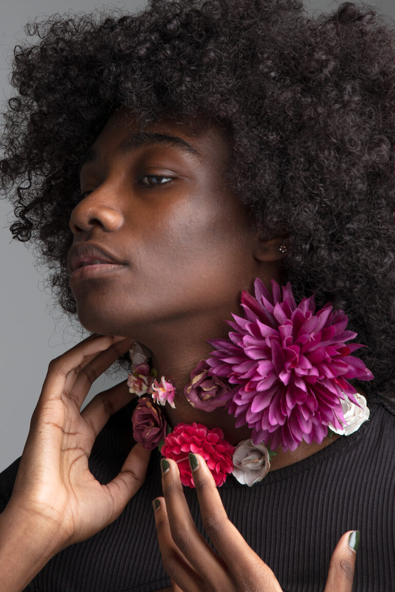 Black woman, with her hair styled in an afro, her left hand is softly touching the side of her neck, her right hand is in front of her. Her gaze is cast downwards. She is wearing a black shirt. Eight flowers of different sizes and species grow on her neck in colours ranging from pink, white, burgundy and purple.