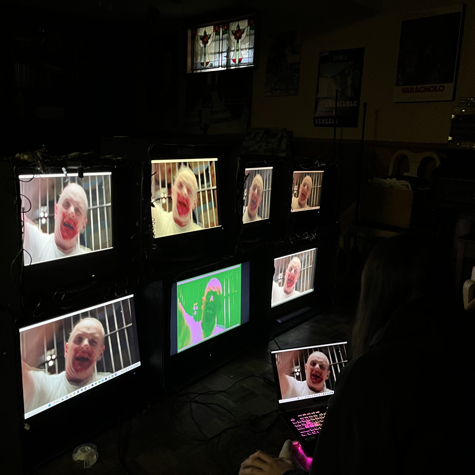 Girl sitting on floor with laptop looking at structure holding 7 monitors displaying a portion of my video work that uses horror movies to represent OCD, this particular still shows Anthony Hopkins in Silence of the Lambs raising his arm to beat someone with a police club