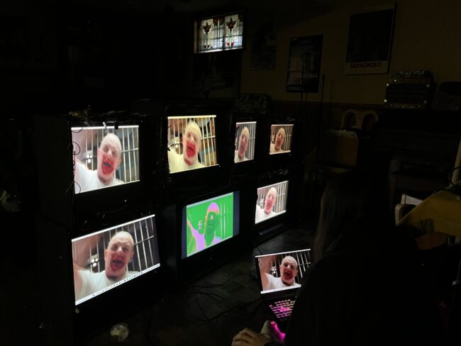 Girl sitting on floor with laptop looking at structure holding 7 monitors displaying a portion of my video work that uses horror movies to represent OCD, this particular still shows Anthony Hopkins in Silence of the Lambs raising his arm to beat someone with a police club