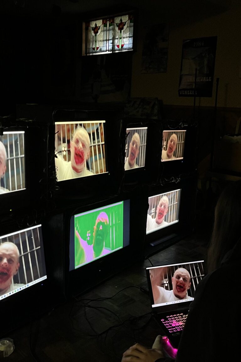 Girl sitting on floor with laptop looking at structure holding 7 monitors displaying a portion of my video work that uses horror movies to represent OCD, this particular still shows Anthony Hopkins in Silence of the Lambs raising his arm to beat someone with a police club