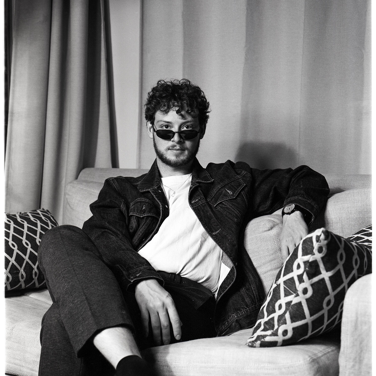 : A black and white image shot on film depicting Jamie, a man in his early 20’s with a beard and short curly hair. Jamie is sitting on a couch in front of a curtain looking directly into the camera. His legs are crossed and he has a straight face. He is wearing a denim jacket with a white t-shirt underneath paired with dark pants as well as thin sunglasses under his eyes.