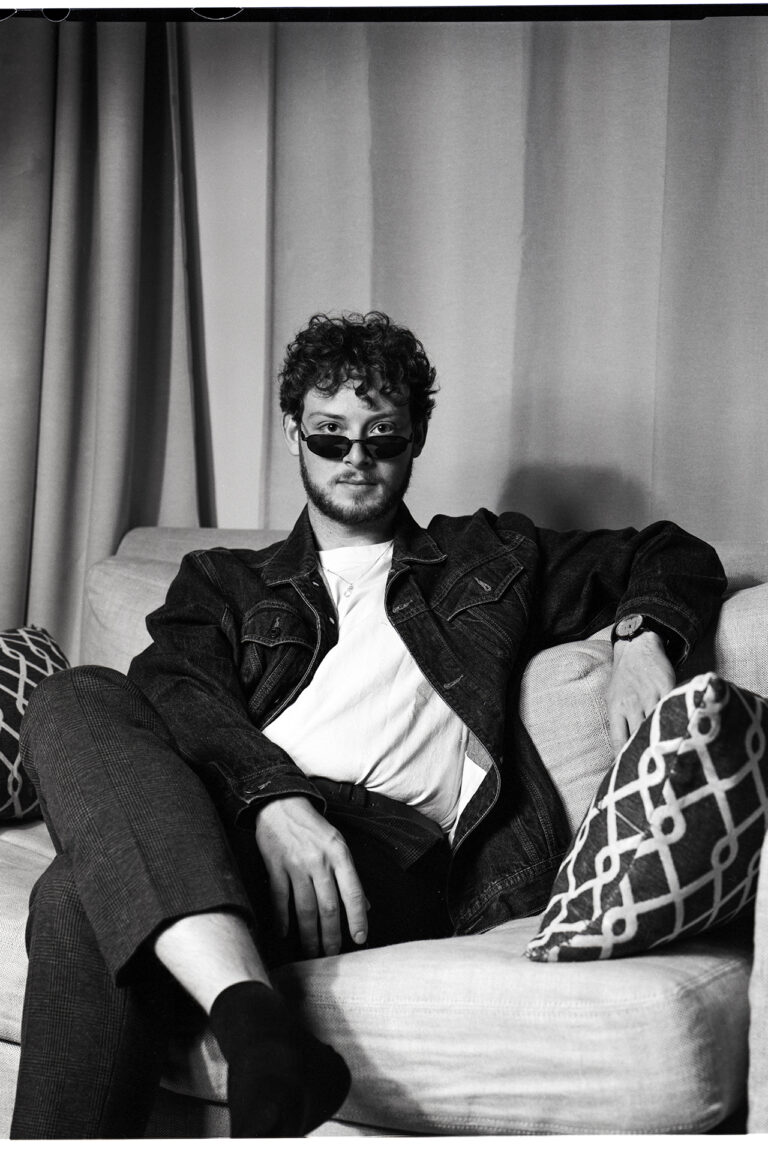 : A black and white image shot on film depicting Jamie, a man in his early 20’s with a beard and short curly hair. Jamie is sitting on a couch in front of a curtain looking directly into the camera. His legs are crossed and he has a straight face. He is wearing a denim jacket with a white t-shirt underneath paired with dark pants as well as thin sunglasses under his eyes.