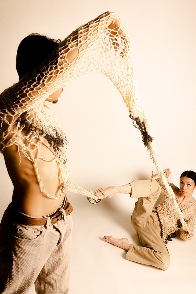 A photograph of two people intertwined by a garment piece. The sleeve of the garment is connected. The first model is standing and on the left side of the frame and is closer to the camera and the second model is sitting on the floor further from the camera.