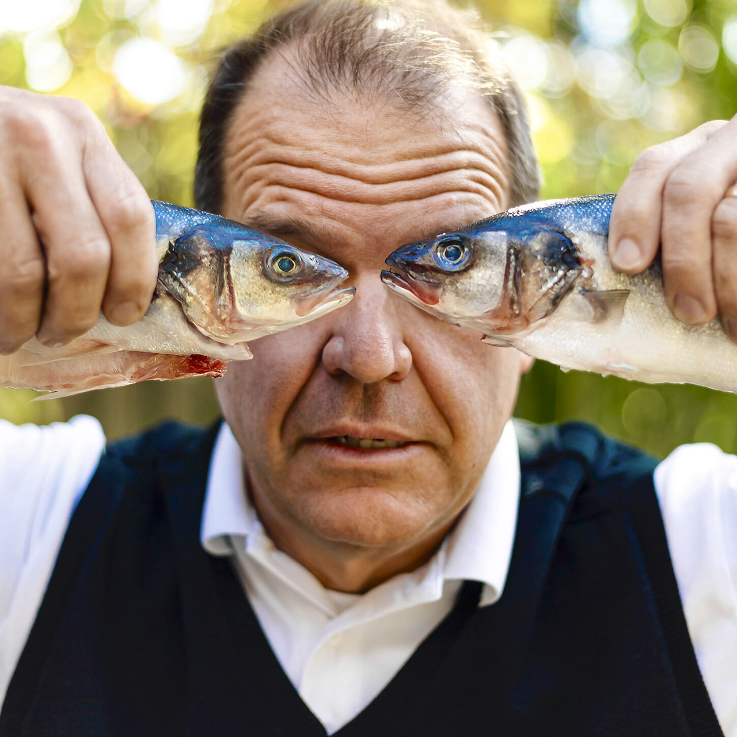 Fish Eyes: A middle-aged, medium-light skinned man is holding a dead fish up to his eyes in each hand. The man is wearing a white collared shirt with a navy blue sweater vest on top, looking directly at the camera.