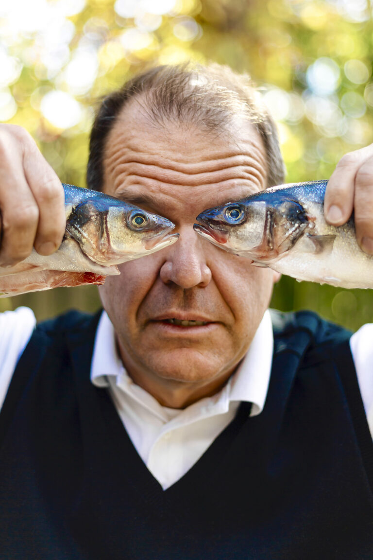 Fish Eyes: A middle-aged, medium-light skinned man is holding a dead fish up to his eyes in each hand. The man is wearing a white collared shirt with a navy blue sweater vest on top, looking directly at the camera.