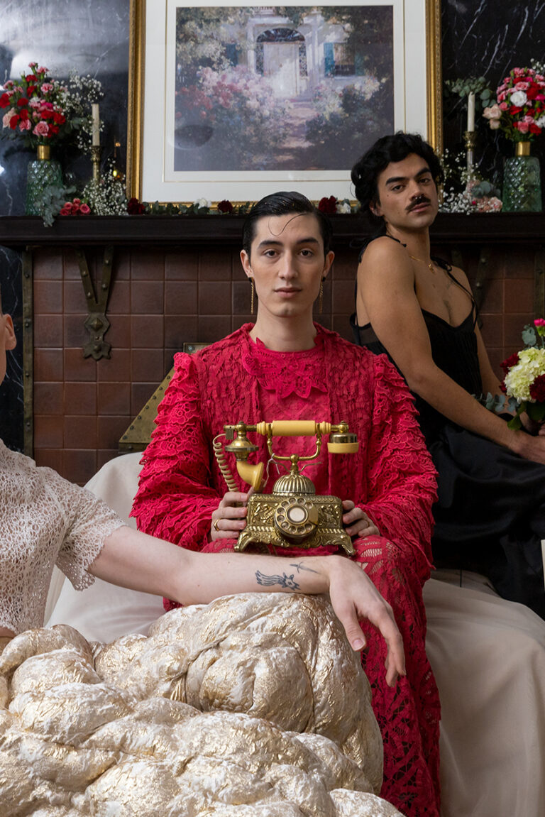 Three models sit in front of a fireplace in a diagonal line. The one in the center holds a gold vintage phone.