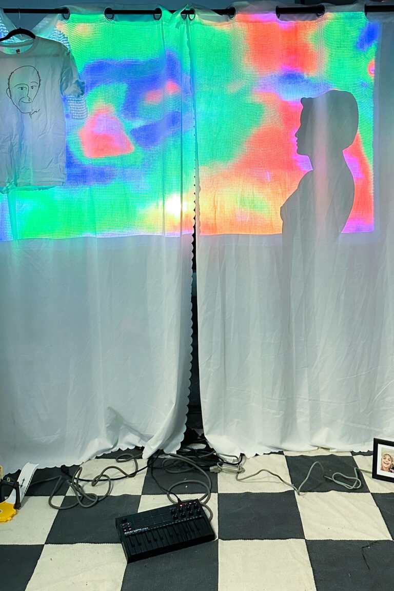 interactive installation with a projection through white curtains. there is a checkered rug with personal items on top (monkey plush, framed photo, heart shaped mirror, midi keyboard, kids toys, exposed wires)