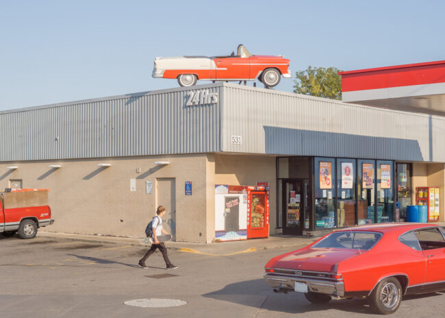 Gas: A skateboarder walks through the scene of a busy gas station, which is visited by and decorated with red cars, both old and new. It's golden hour, and the sky is a bright blue. The centre point of the image is an old fake plastic red car on top of the gas station, placed as decoration.