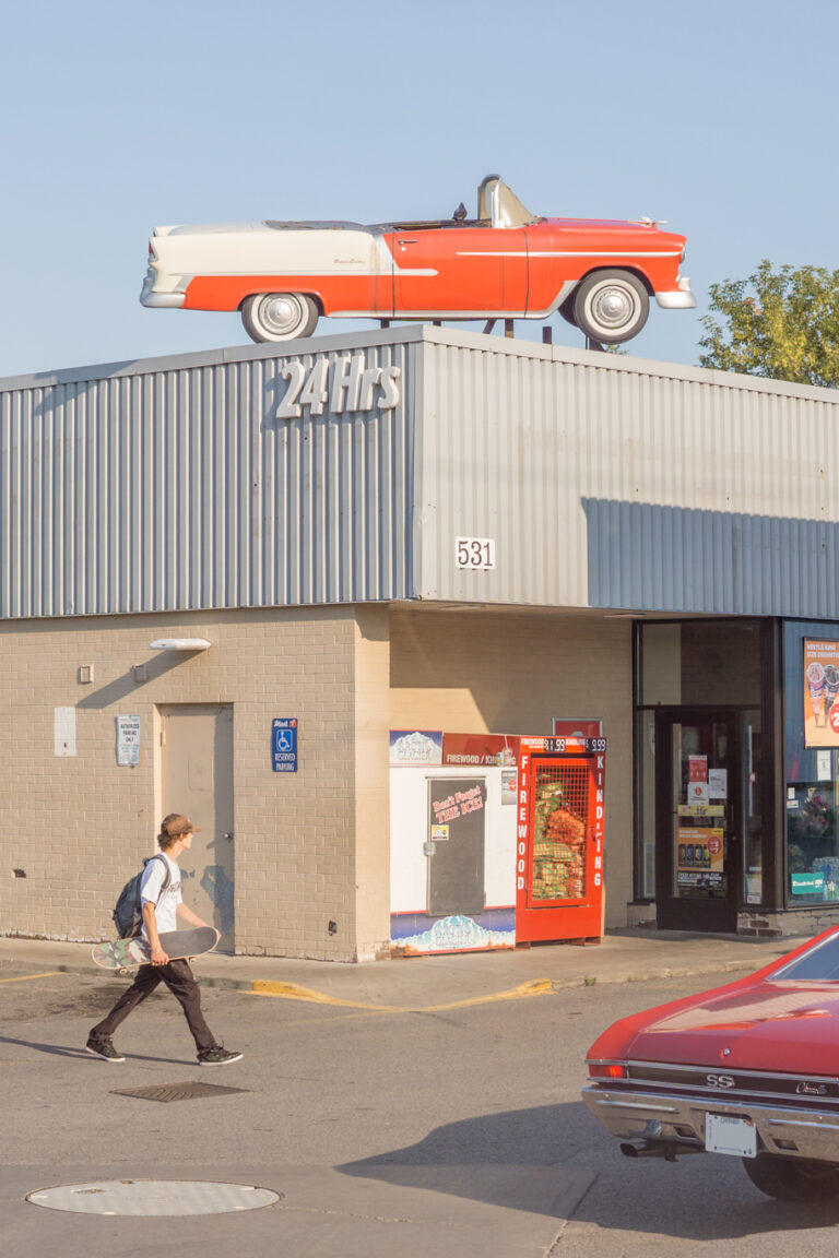 Gas: A skateboarder walks through the scene of a busy gas station, which is visited by and decorated with red cars, both old and new. It's golden hour, and the sky is a bright blue. The centre point of the image is an old fake plastic red car on top of the gas station, placed as decoration.