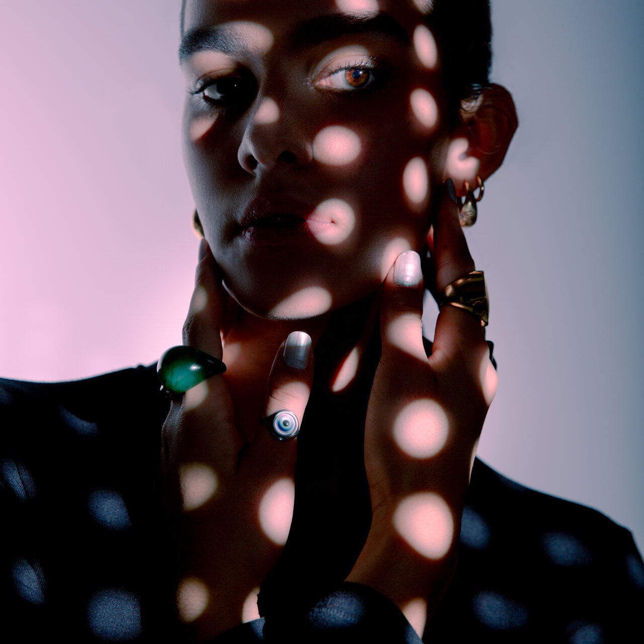 Adriana is posing with a circular pattern of lights hitting her face and upper body. She's wearing a black full sleeve top. Her accessories and eyes shine through in the light against a gradient background.