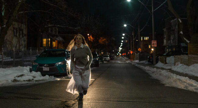 A young, blonde, white woman runs in the middle of a residential street towards the camera on the left side of the image. She is wearing a grey sweater with white collar and a car light behind her illuminates her long white skirt. The image is cool-toned and tinted green.