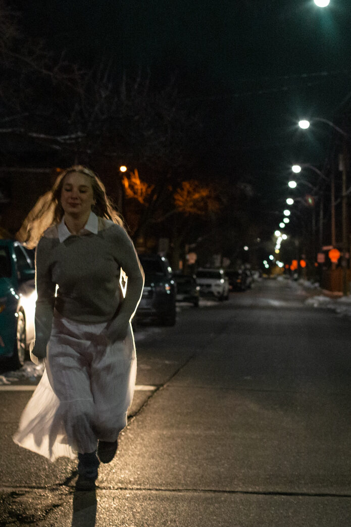 A young, blonde, white woman runs in the middle of a residential street towards the camera on the left side of the image. She is wearing a grey sweater with white collar and a car light behind her illuminates her long white skirt. The image is cool-toned and tinted green.