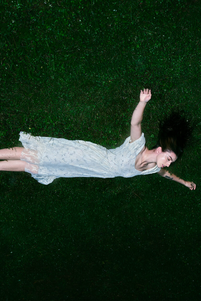 Queer - A young woman in a white dress is lying on the ground, placed at the top of the image. The vibrant green grass is wet, and her white dress stands out against the darkness. She looks to be both falling and held in suspension. Her eyes are shut and her limbs are splayed out around her, with her arms forming a T and her legs stretching straight out.