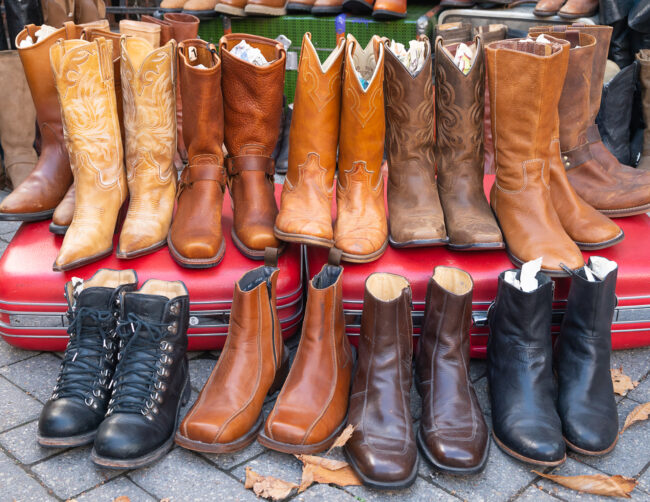 Worn out pairs of brown and black leather cowboy boots. They are displayed outside on the ground, propped up on a red vintage suitcase.
