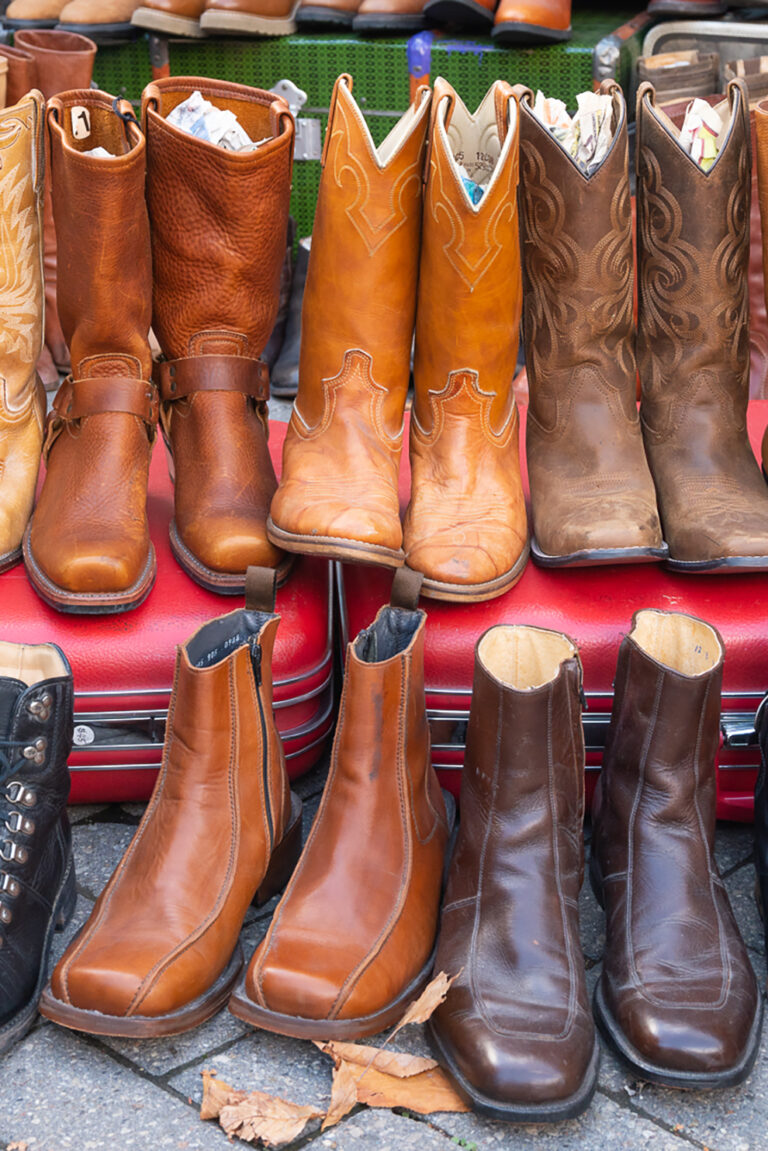 Worn out pairs of brown and black leather cowboy boots. They are displayed outside on the ground, propped up on a red vintage suitcase.