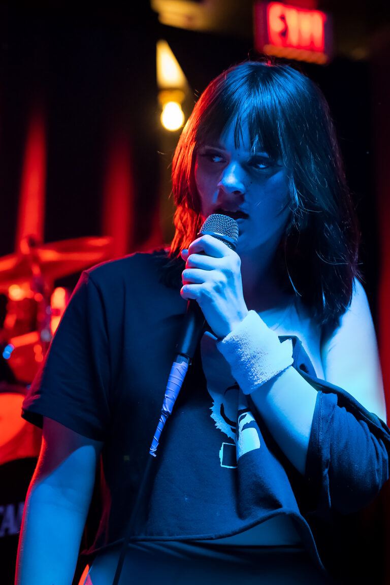 Redd, lead vocalist for band "Electrokat", is posed singing mid-performance wearing 80s style workout gear. She looks off in thought in front of a drumset, featured in contrasting red and blue lights.
